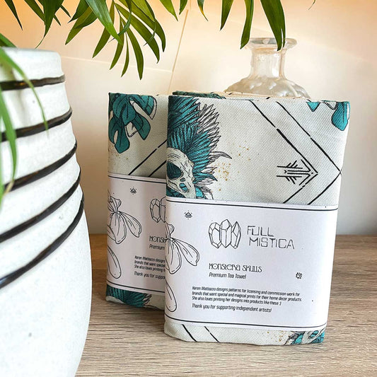 Two tea towels standing on wooden table, leaning on glass bottle. Plant pot close up to the left, leaves falling from above. Designs on towels are tropical leaves in teal with bird skull, runes and lines in black.